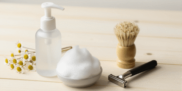 Espuma de barbear caseira num frasco espumador com pincel, aparelho e flores de camomila ao lado