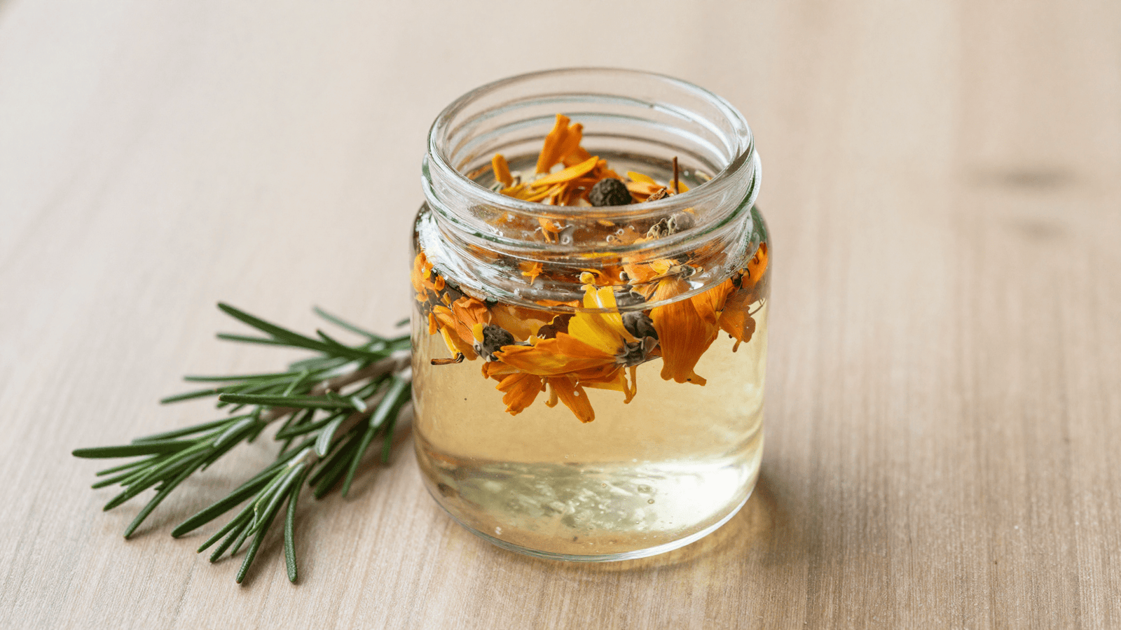 Flor de calendula triturada macerando na glicerina dentro de um pote de vidro com alecrim ao lado