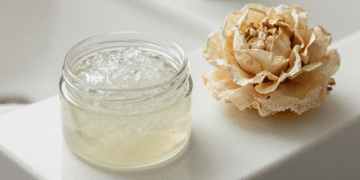 Pote de gel corporal esfoliante transparente com grânulos ao lado de uma bucha vegetal e flor seca