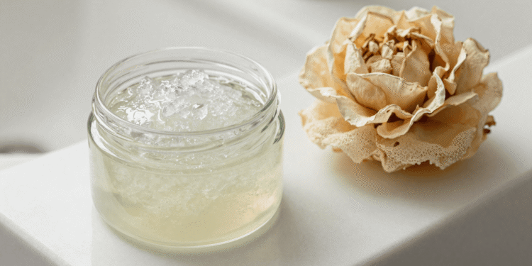 Pote de gel corporal esfoliante transparente com grânulos ao lado de uma bucha vegetal e flor seca