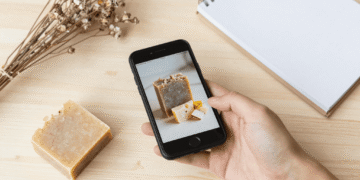 Bancada de artesã com celular mostrando foto de sabonete, barras artesanais e flores secas sobre mesa clara